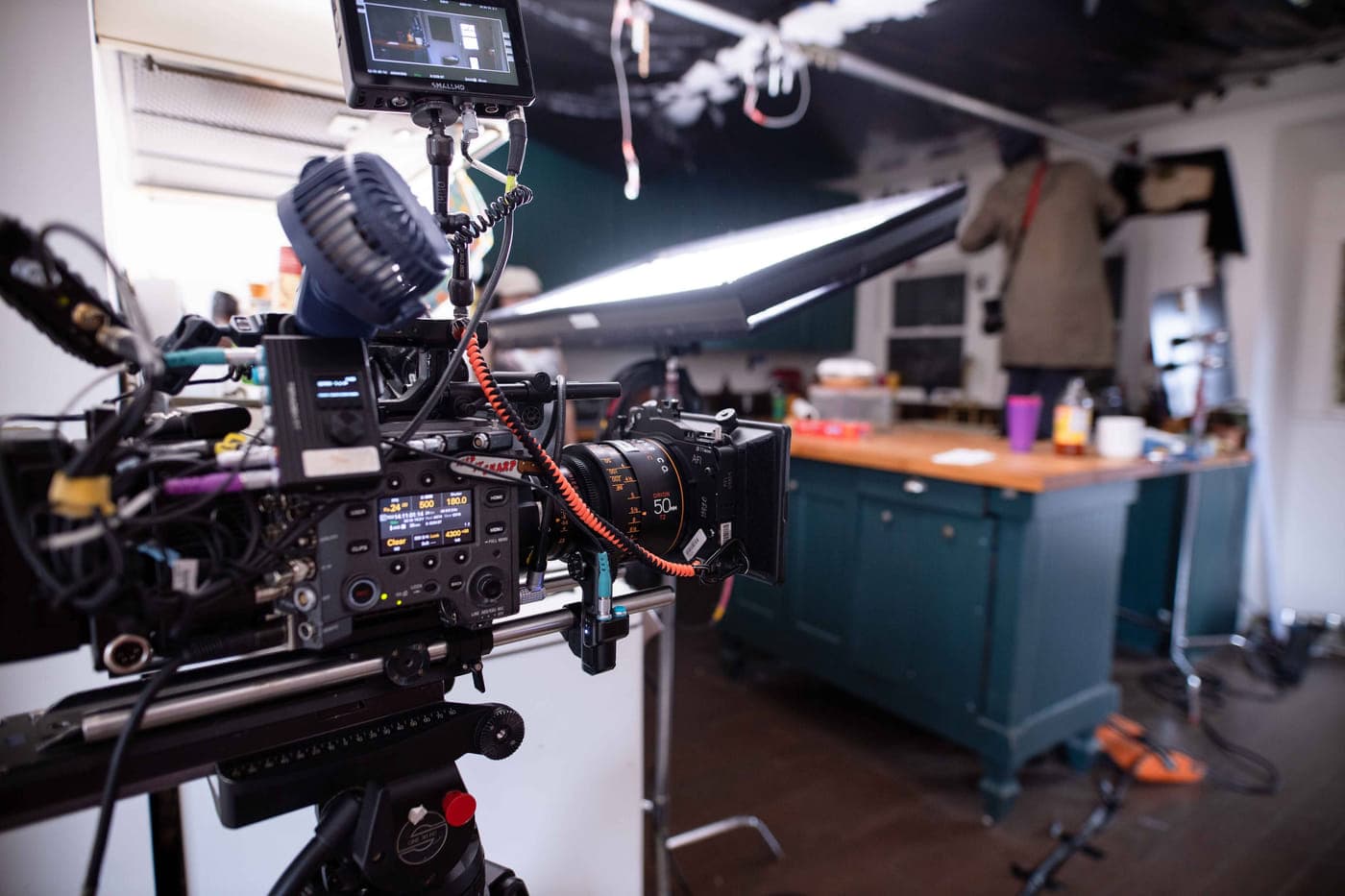 A cinema camera built on a tripod with a prime lens and matte box, facing a lit interior set